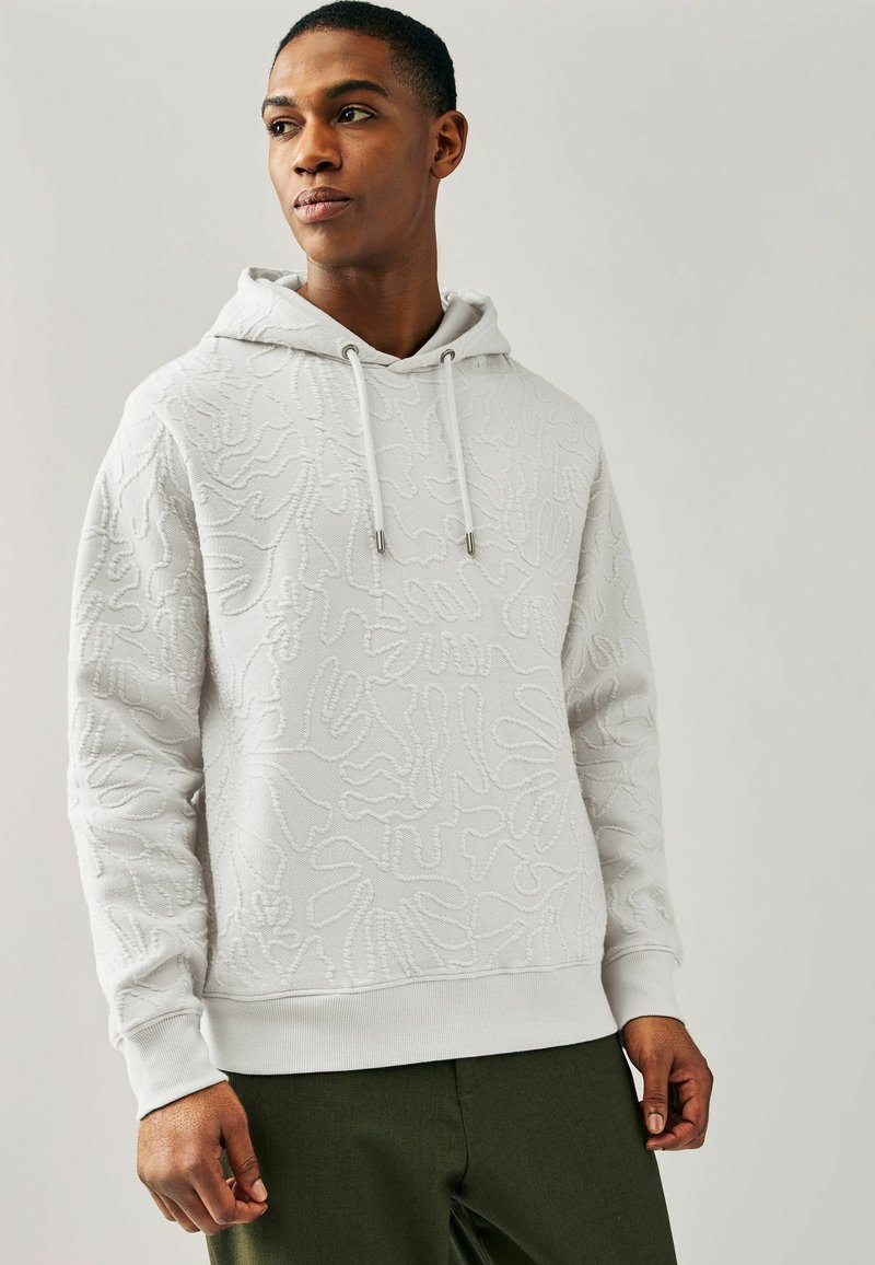 Sudadera blanca con patrón texturizado, capucha con cordones, puños y dobladillo de canalé. Combinada con pantalones verde oliva. Fondo claro.