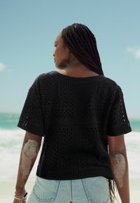 Sort strikket top med korte ærmer, der har et hulmønster. Båret med lyseblå denimshorts, sandfarvet hud på armen, havbaggrund.