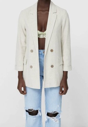 Personne à la peau foncée portant un blazer beige croisé, un bralette en dentelle vert clair et un jean bleu clair déchiré.