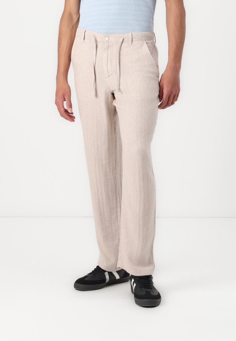 Lys beige linbukser med lige skæring, snøre i taljen og sidelommer, parret med sorte sneakers.