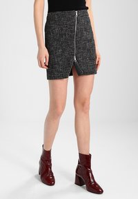 Mini-jupe en tweed noir avec fermeture éclair frontale et fente latérale. Elle présente un tissu texturé et un accent en similicuir au niveau de la fermeture éclair.