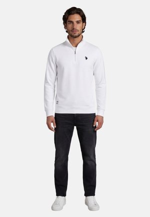 Mann trägt weiße Sweatshirt mit Viertelreißverschluss, schwarze Jeans und weiße Turnschuhe, steht vor einem schlichten hellen Hintergrund.