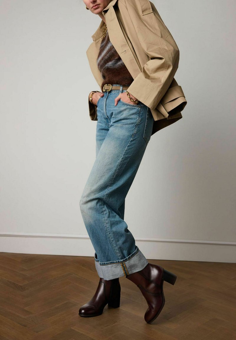 Blue denim jeans with rolled cuffs, brown heeled boots, and a tan oversized jacket. Textured brown sweater underneath.