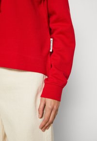 Sudadera de algodón roja con puños acanalados, que presenta una pequeña etiqueta de logo en la manga. Combinada con pantalones beige claros.