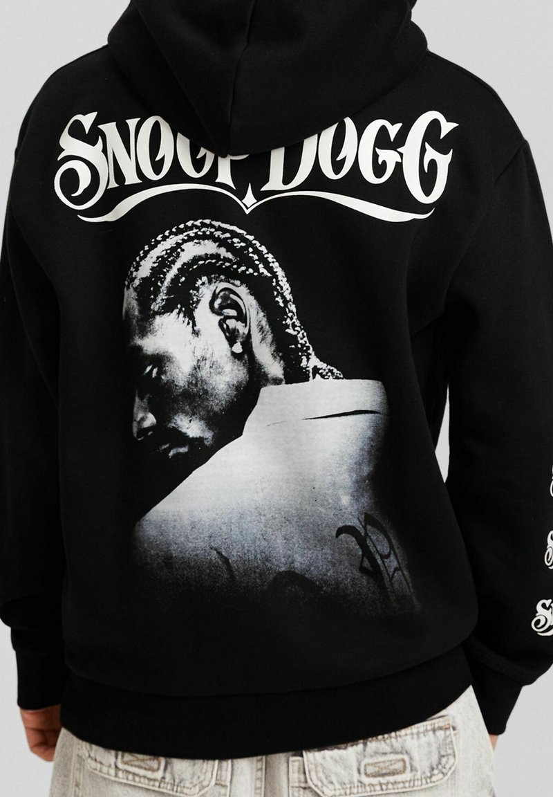 Persona che indossa un hoodie nero con la scritta bianca "Snoop Dogg" e un ritratto in bianco e nero di profilo stampato sul retro.