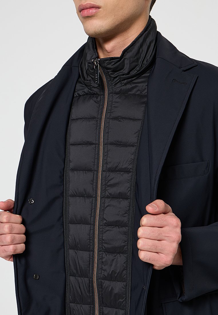 Schwarzige Puffer-Weste mit vertikaler Steppung, getragen unter einer dunklen Jacke; sie hat einen hohen Kragen und einen Frontreißverschluss mit einem bronzefarbenen Zieher.