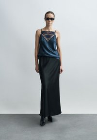 Marineblaue Satin-Camisole mit Spitzenbesatz, kombiniert mit einem bodenlangen schwarzen Rock. Das Model trägt schwarze Sonnenbrillen und elegante schwarze Stiefel.