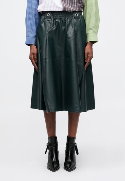 Dunkelgrüner Kunstleder-Midi-Rock mit elastischem Bund, Ösen-Details und glatter Textur, kombiniert mit schwarzen Ankle-Boots.
