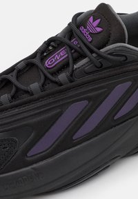 Svart Adidas sneakers med lila accenter, svarta snören, "adiPRENE" sula och "Ozweego" etikett på tungan.