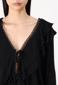Svart blus med djup V-ringning, texturerad tyg och rynkade detaljer, accentuerad med intrikata spetskanter och en knytdetail vid halsen.