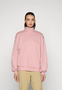 Pullover con cerniera rosa chiaro, colletto alto, spalle basse e polsini a costine. Presenta un piccolo logo bianco sul petto. Tessuto morbido.