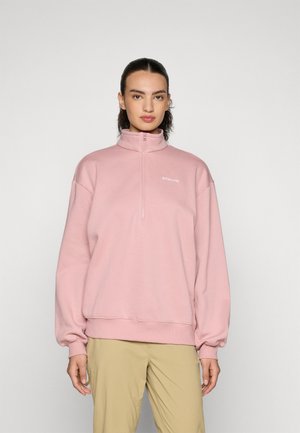 Light pink zip-up pullover with a high collar, dropped shoulders, and ribbed cuffs. Features a small white logo on the chest. Soft fabric texture.