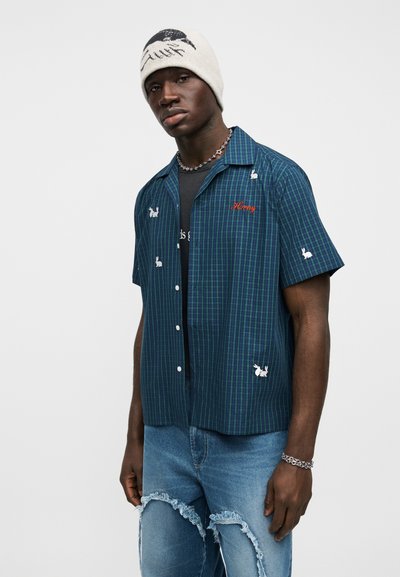 Άνδρας που φοράει μπλε καρό κοντομάνικο πουκάμισο με λευκές εκτυπώσεις λαγουδάκια, γκρι σκουφάκι, κολιέ με αστέρι, σκισμένο μπλε τζιν και ασημένιο βραχιόλι.