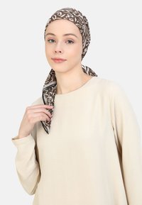 Brown patterned scarf made of lightweight fabric, featuring white floral designs. Worn draped around the head and shoulders over a beige top.