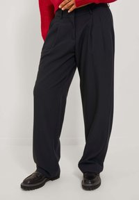 Svarta plisserade byxor med en lös passform. Tillverkade av ett mjukt tyg, med hög midja och sidofickor. Matchat med svarta skor.