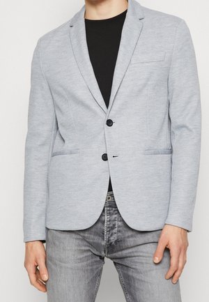 Mężczyzna w jasnoszarej, teksturowanej marynarce z dwoma czarnymi guzikami, czarnej koszuli i szarych dżinsach, stojący ze swobodnie opuszczonymi rękami.