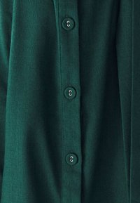 Temno zelena srajca z gumbom, izdelana iz teksturirane tkanine, s petimi usklajenimi gumbi vzdolž sprednjega dela in ovratnikom.