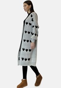 Długi szary dziany kardigan z czarnymi wzorami w kształcie serc, ściągaczami na rękawach i na dole. Noszony na czarnym topie, zestawiony z czarnymi legginsami i brązowymi botkami.