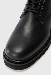 Botas de cuero negro con puntera redondeada, superficie texturizada, costuras en contraste y ojales para los cordones. Suela de goma gruesa.