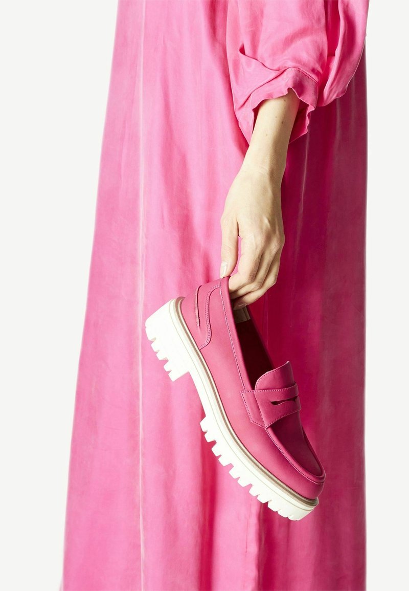 Chaussure en cuir rose avec une semelle blanche épaisse, dotée d'un design à enfiler et de lignes d'accent horizontales. Présentée à la main sur un fond de robe rose.