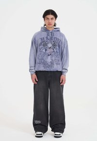 Joven con cabello oscuro de longitud media que lleva una sudadera azul oversized con un gráfico de una cruz y calaveras, pantalones negros amplios y zapatillas en blanco y negro.