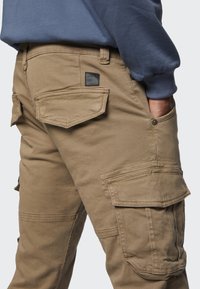 Beige Cargo-Hosen mit mehreren Klapptaschen, einem strukturierten Stoff und einem Label auf der Rückseite, kombiniert mit einem blauen Sweatshirt.