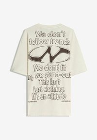 Bež oversized majica s velikim crnim grafikom i natpisom: "Ne slijedimo trendove. Ne uklapamo se, već se ističemo. Ovo nije samo odjeća. To je stav."