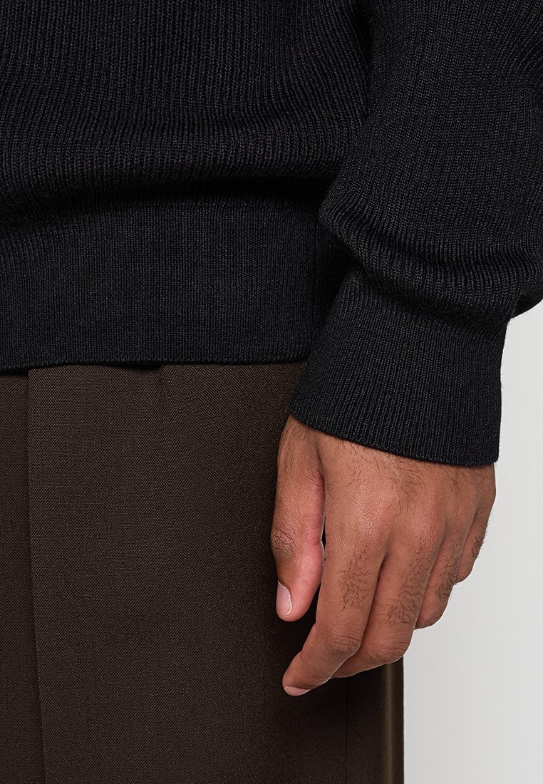 Schwarzer gerippter Pullover mit strukturiertem Strick und engen Bündchen, kombiniert mit braunen Stoffhosen. Nahaufnahme einer Hand, die auf dem Stoff ruht.