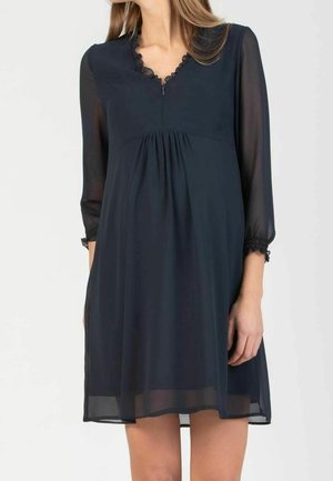 Frau trägt ein locker sitzendes schwarzes Kleid mit transparenten langen Ärmeln, geraffter Taille und geschwungenem Ausschnitt, steht vor einem schlichten Hintergrund.