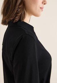 Camisa negra con cuello, de textura suave, con un diseño ajustado y mangas cortas, mostrando un perfil lateral de una persona.