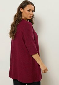 Tunika a maniche lunghe color bordeaux con vestibilità rilassata e tessuto strutturato. Presenta spacchi laterali e scollo rotondo.