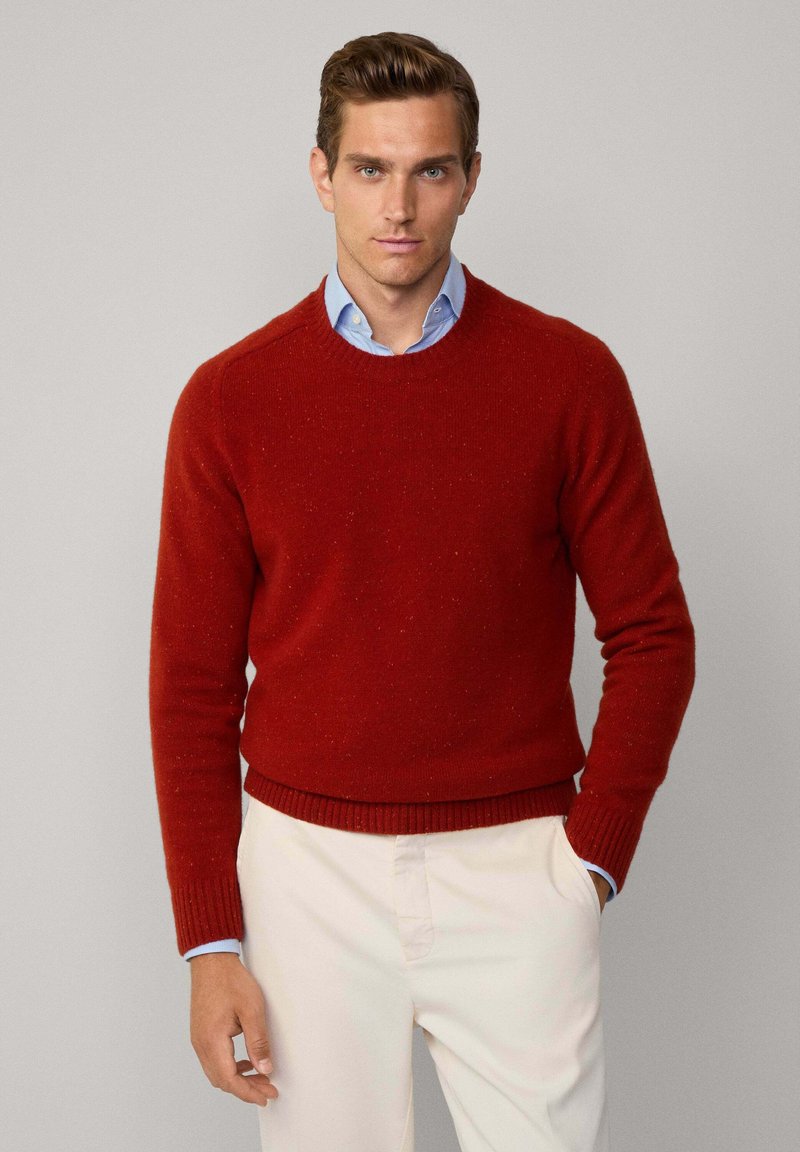 Roter Strickpullover mit Rundhalsausschnitt und gerippten Bündchen. Darüber wird ein hellblaues Kragenshirt getragen, kombiniert mit hellen Hosen.