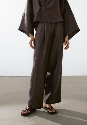 Personne portant un pantalon large ample marron foncé et un haut assorti à larges manches, associé à des sandales noires avec anneau d’orteil.