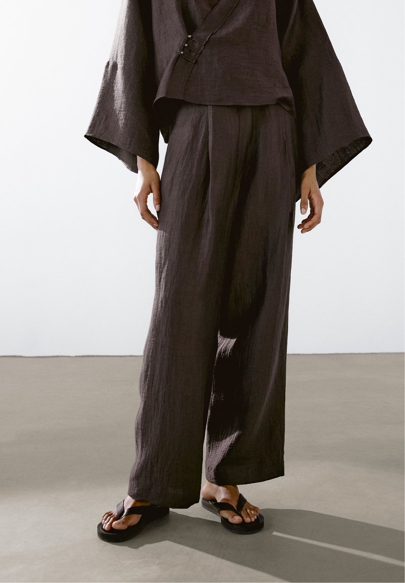 Personne portant un pantalon large ample marron foncé et un haut assorti à larges manches, associé à des sandales noires avec anneau d’orteil.