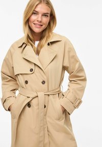 Trench-coat kaki à double boutonnage, grand col et taille ceinturée. Comprend deux poches avant et une texture lisse.