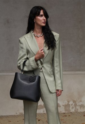 Woman with long black hair in sage green suit holds large black handbag, wearing chunky silver necklace and earrings, standing against textured wall.