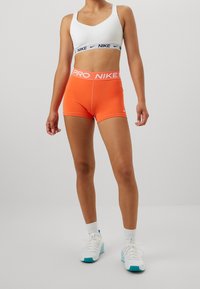 Biały sportowy biustonosz Nike z czarnymi akcentami; pomarańczowe dopasowane szorty z logo "PRO Nike"; zestawione z białymi trampkami z niebieskimi akcentami.