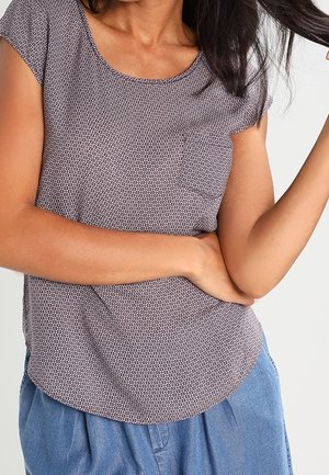 Vrouw met donker haar draagt een kortermelig top met patroon en blauwe broek, met één arm gebogen over haar taille en de andere omhoog.