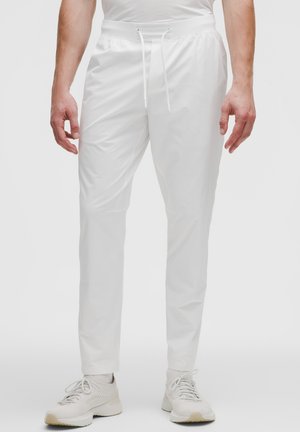 Hombre con pantalones jogger blancos ajustados con cintura de cordón y zapatillas blancas, de pie contra un fondo liso y claro.