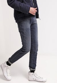 Persona con chaqueta azul oscura, vaqueros desgastados azul claro arremangados, calcetines negros y zapatillas blancas de caña alta caminando con la pierna izquierda hacia delante.