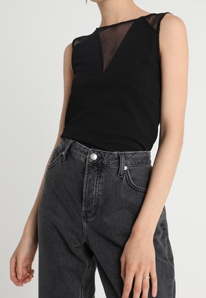 Czarny top bez rękawów z przezroczystą siateczkową wstawką na górnej części klatki piersiowej, zestawiony z wysokim stanem wyblakłych czarnych dżinsów na stojącej osobie.