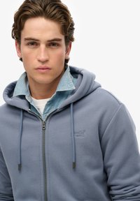 Blauwe zip-up hoodie van zacht materiaal, met een voorrits, trekkoorden en een logo op de borst. Gelaagd over een spijkerhemden.