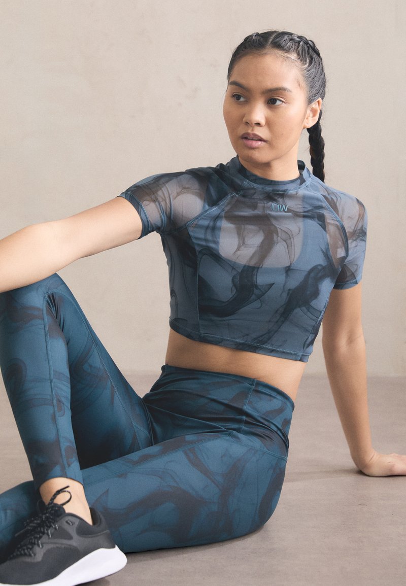 Crop top de sport et leggings en bleu sarcelle avec un motif abstrait. Le matériau semble lisse et extensible, avec un design ajusté et des manches courtes.