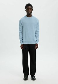 Svjetloplavi pleteni pulover s dugim rukavima i rebrastim manžetama, kombiniran s crnim krojenim hlačama i crnim cipelama. Jednostavan, čist dizajn.