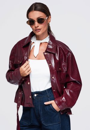 Femme portant une veste rouge foncé brillante avec un motif crocodile, un haut blanc avec une cravate, un jean bleu foncé et des lunettes de soleil rondes, posant avec une main dans la poche.