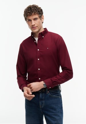 Junger Mann mit lockigem Haar, der ein dunkelrotes Hemd mit Knöpfen und blaue Jeans trägt, steht und richtet seinen Ärmel vor einem einfarbigen Hintergrund.