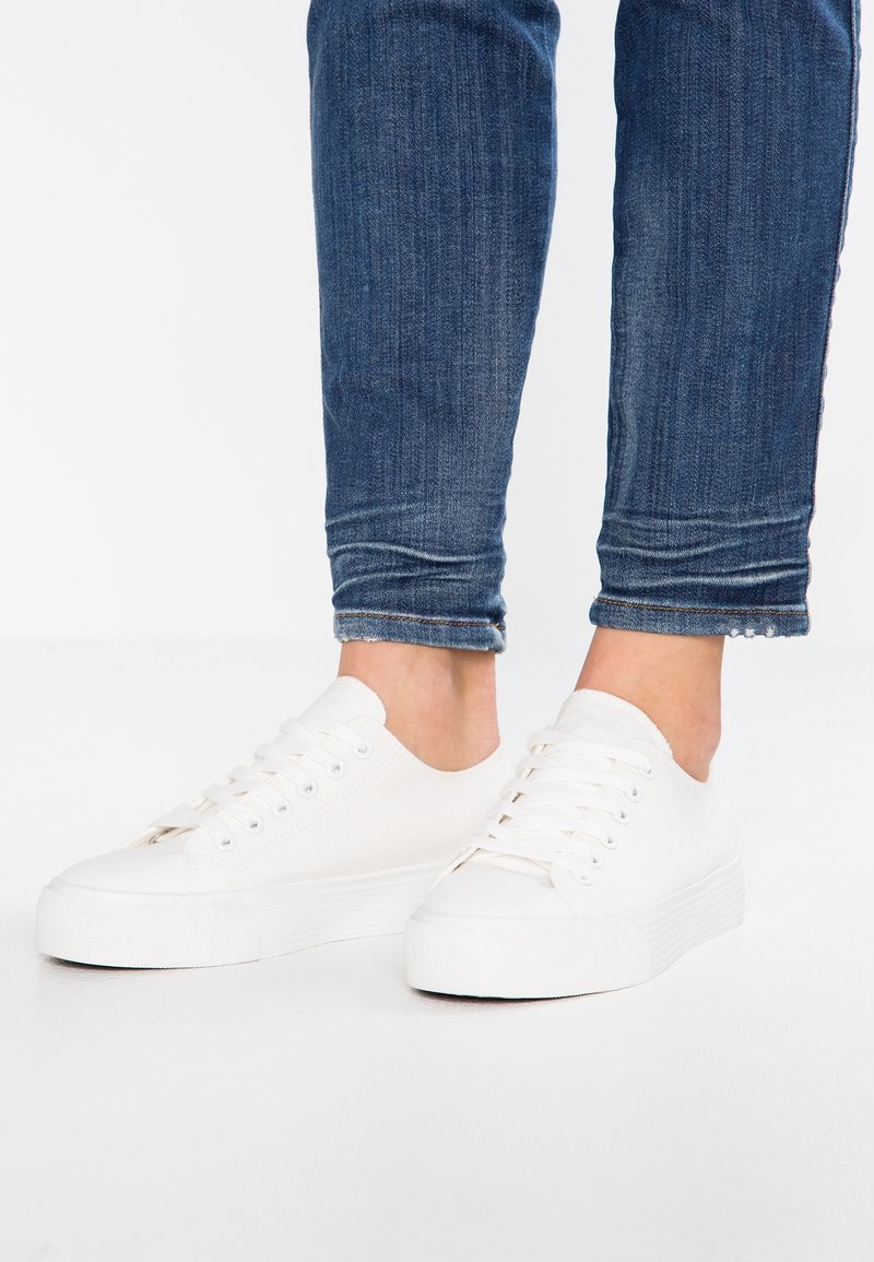 Personne portant un jean bleu retroussé et des baskets basses blanches à lacets, debout sur un sol blanc.