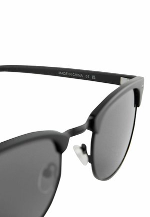 Černé sluneční brýle s matnými rámy a šedými skly. Mezi jejich vlastnosti patří plochý design, výrazná obroučka obočí a kovové nosní podložky.