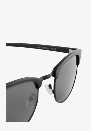 Juodos saulės akiniai su matiniais rėmeliais ir pilkais objektyvais. Savybės: plokščias dizainas, išreikštas antakio linija ir metaliniai nosies kaitikliai.