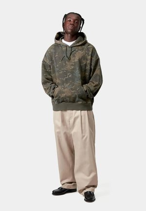 Mann steht, trägt grünen Camouflage-Hoodie, lockere beige Hose und schwarze Schuhe vor einem einfarbigen hellen Hintergrund.